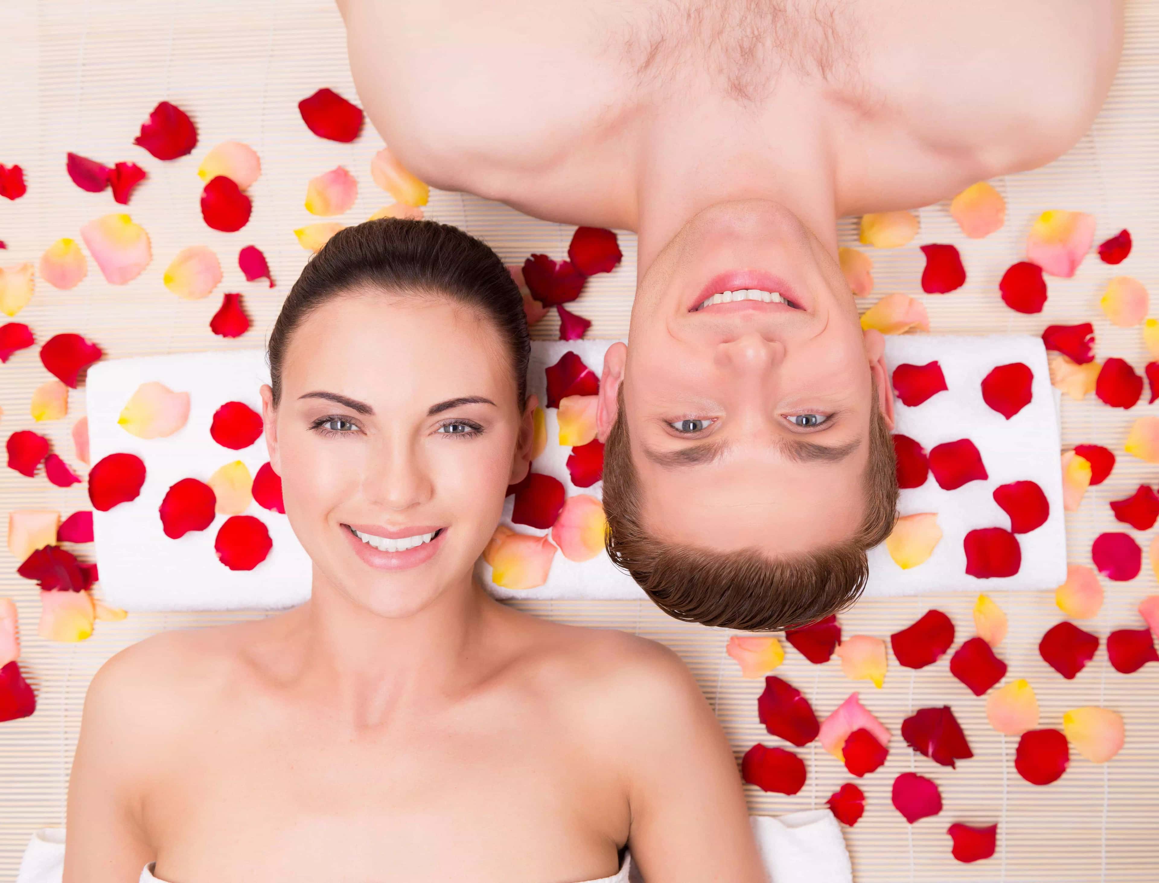 Rostos de homem e mulher. Eles estão deitados sorrindo para a foto. Ao redor de suas cabeças têm pétalas de rosas. Ambos estão em lados opostos para que o rosto de cada um fique lado a lado.