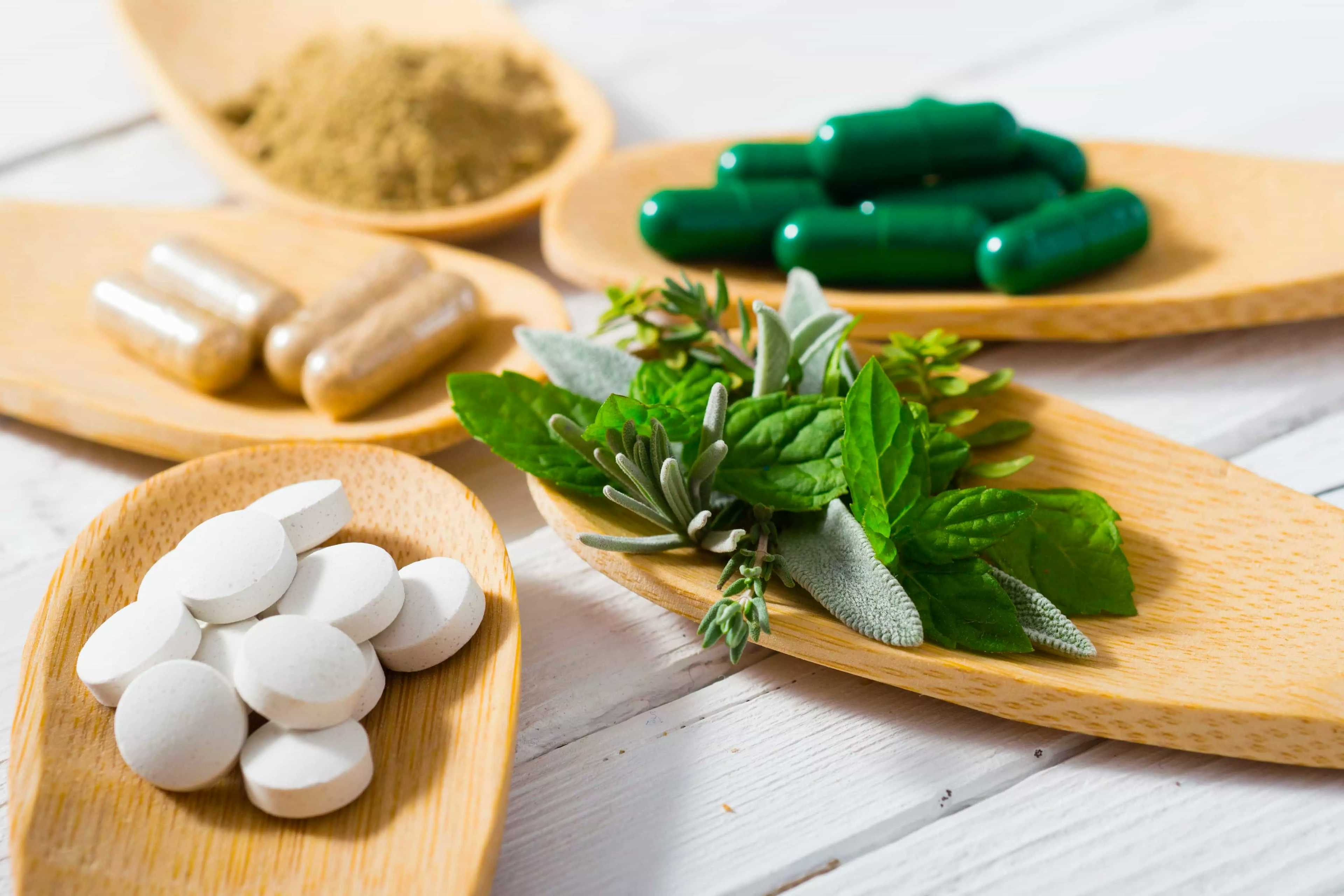 Três tipos de medicamentos naturais sobre um recipiente de madeira, além de um em textura de pó. Também há folhas verdes sobre um outro recipiente.